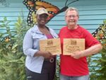 Two people holding wooden plaques awards in front of a flowerbed and mural painted with butterflies.